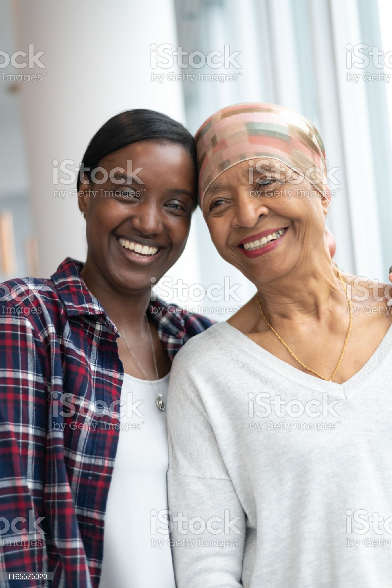 A senior African American woman with cancer stands near a large wall of windows. She is embracing her adult daughter. They are smiling while resting their foreheads against each other. The women are full of hope for the future.