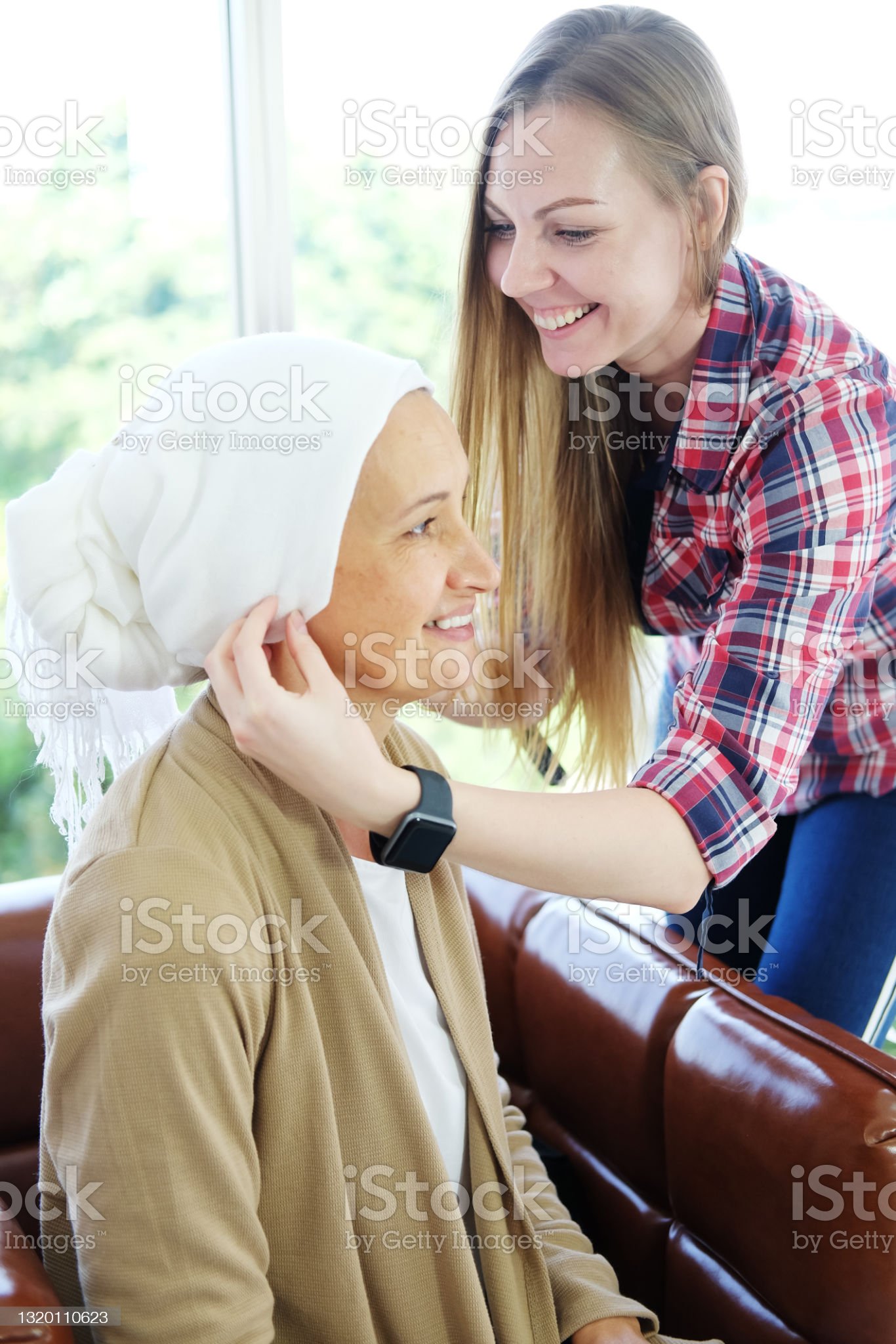 Smiling Caucasian young daughter is hugging and caring her Elderly Mother in white headscarf after chemotherapy because she is suffering from cancer or Leukemia patient. Focus on young woman