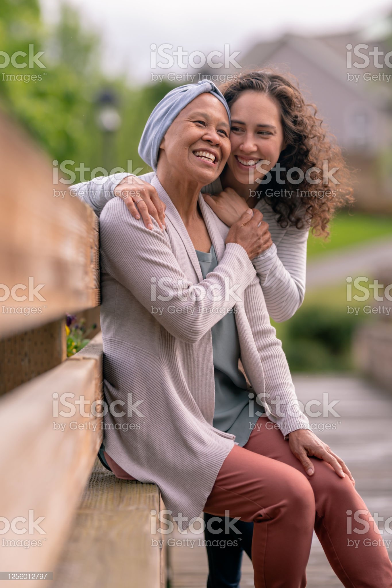 A senior woman with cancer is embraced and comforted by her adult daughter as they stand outside on a spring evening. Both have serious but content expressions as they savor their time together. Their hands are clasped together.